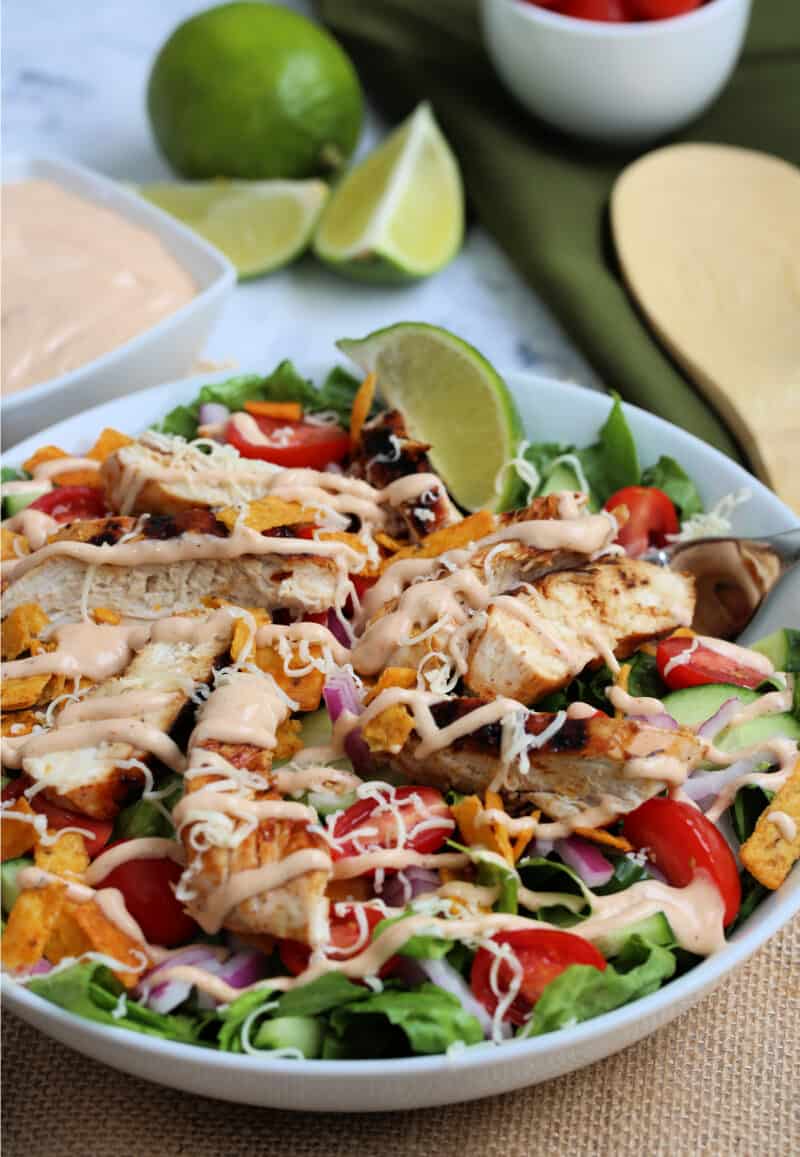 grilled chicken and vegetables in a salad in a bowl with dressing on it with a wooden spoon, dressing and lime wedges on the table