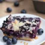 a blueberry pie bar dessert on a white plate surrounded with fresh blueberries.