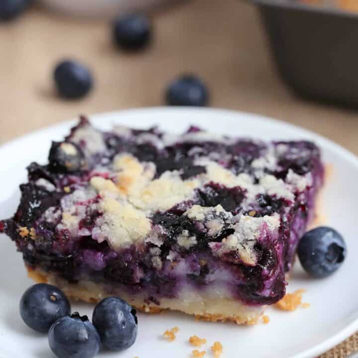 a blueberry pie bar dessert on a white plate surrounded with fresh blueberries
