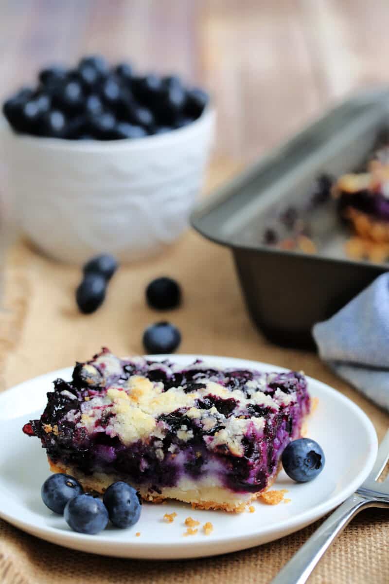 a slice of blueberry square on a white plate with berries surrounding it