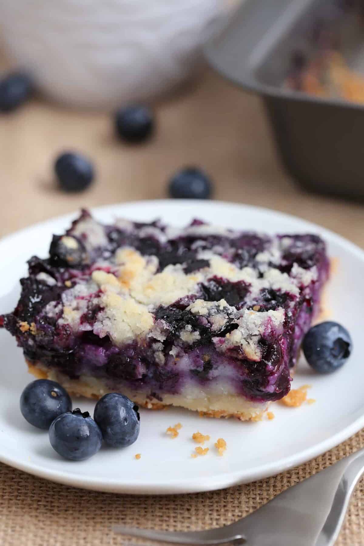 a blueberry pie bar dessert on a white plate surrounded with fresh blueberries.