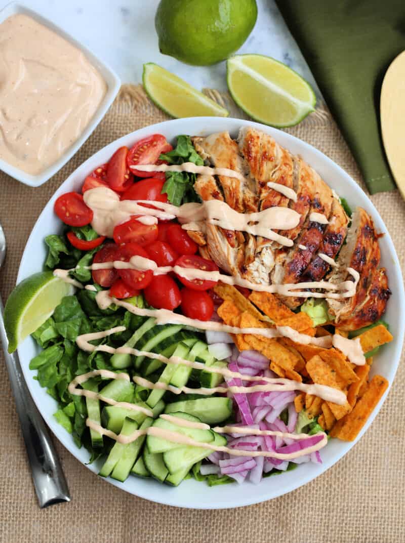 vegetables and bbq chicken slices in a bowl drizzled with dressing with lime wedges and a dish of dressing