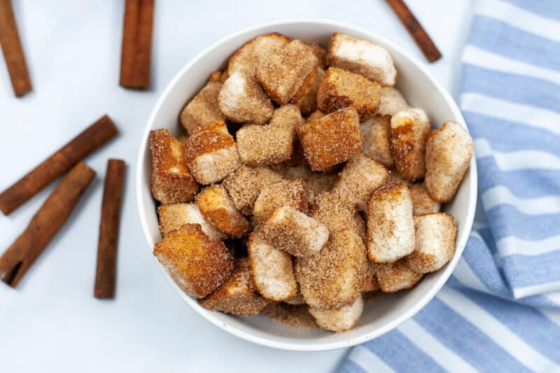overhead photo of a white bowl filled with churro pieces with sticks of cinnamon beside it