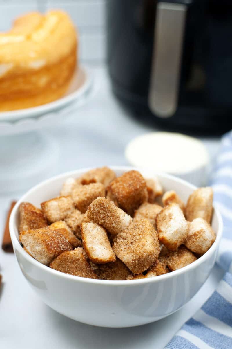 white bowl filled with churro dessert pieces on a table with an air fryer and cake behind it