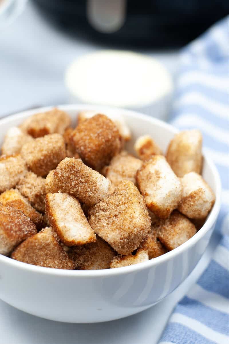 a white bowl of churro dessert pieces beside a blue and white cloth with an air fryer in the background