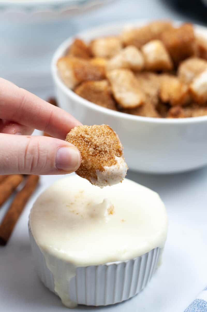 fingers holding a piece of churro over a bowl of white cream cheese frosting