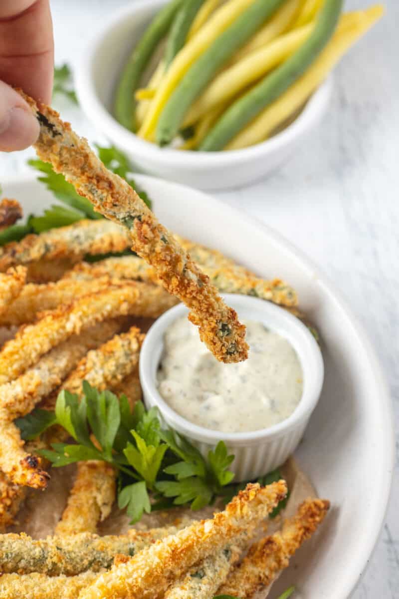 coated air fried green bean held over a white bowl of dip
