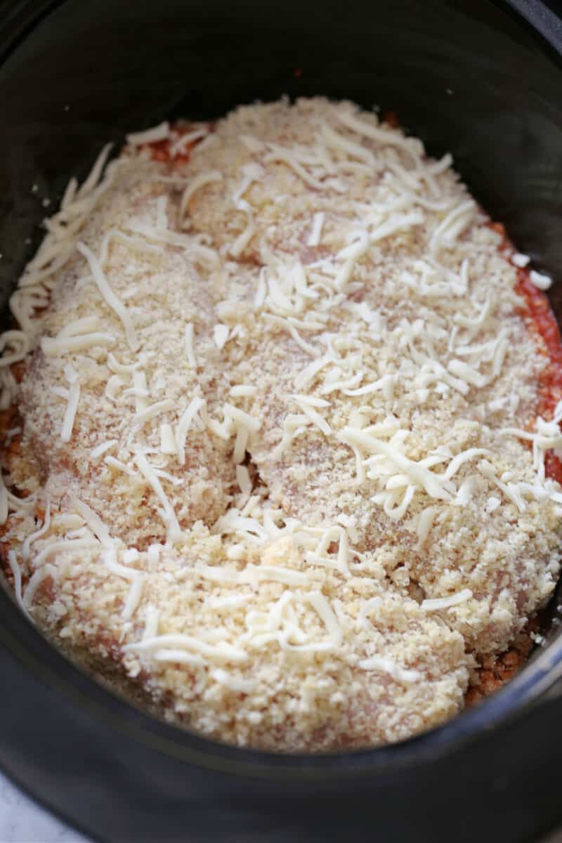 breaded chicken breasts on marinara in a black slow cooker topped with shredded cheese