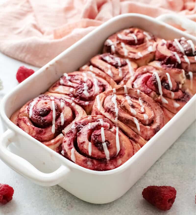 a white dish with raspberry cinnamon rolls inside with raspberries beside it on the table