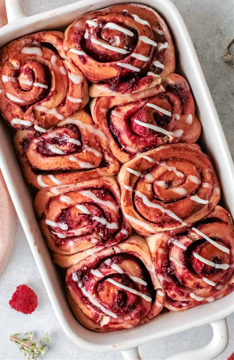 a white dish with raspberry sweet rolls inside and cream cheese drizzle on top