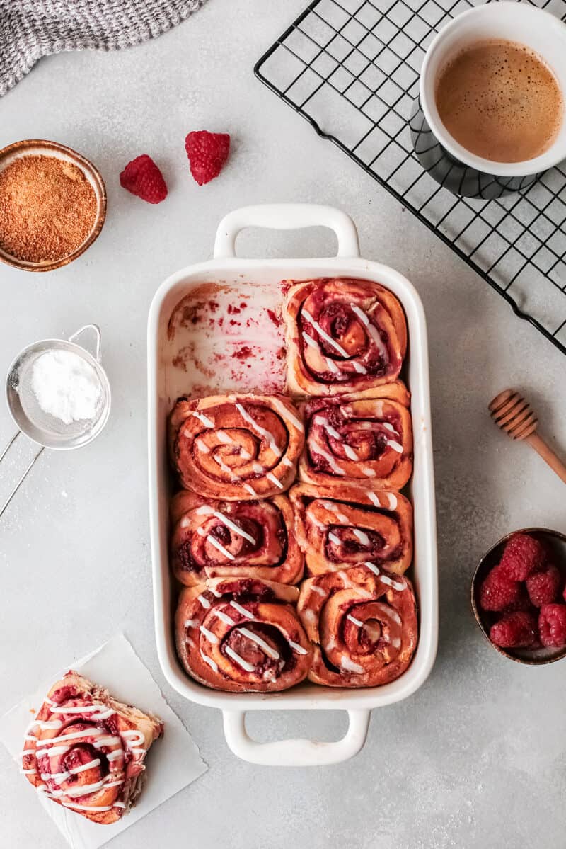 white baking dish with a raspberry sweet roll missing, on a table with ingredients beside it