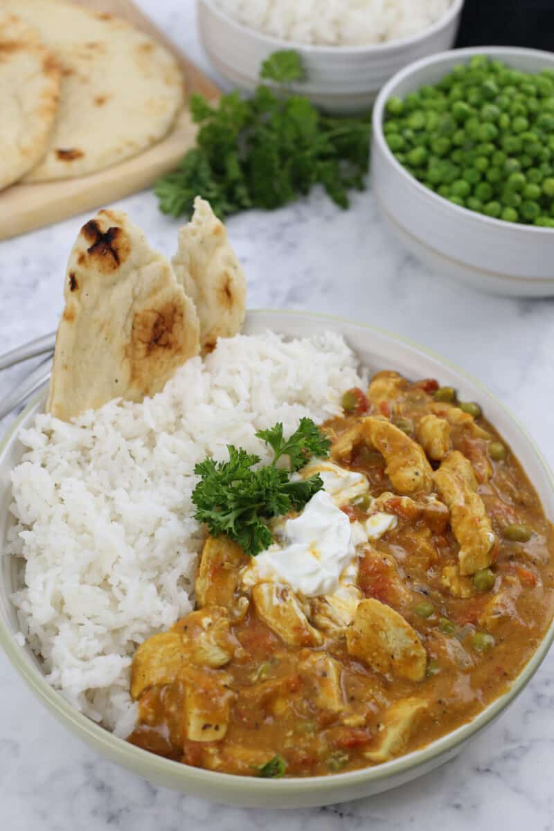 a bowl of chicken curry, rice and naan bread on a table with a bowl of peas