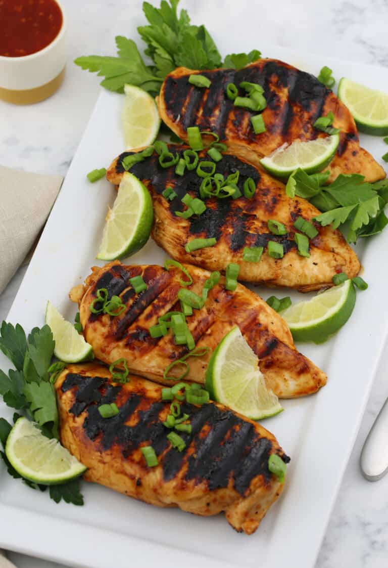 a top view of 4 grilled chicken breasts on a white plate with parsley and lime wedges around them