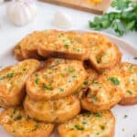 cooked slices of garlic bread with fresh parsley on top on a white plate beside cloves of garlic and shredded cheese