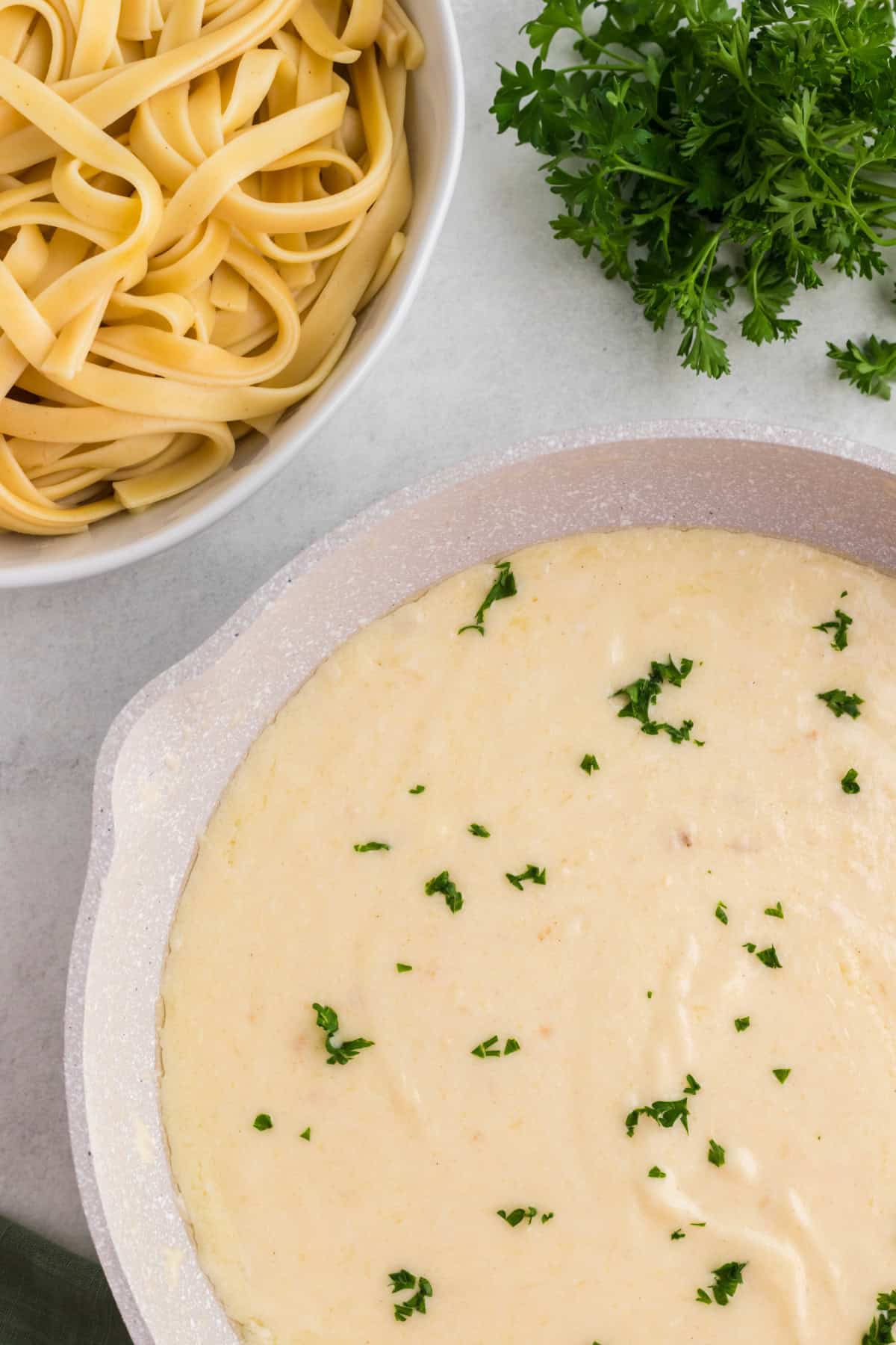 a pan of white sauce with fresh parsley in it beside a bowl of cooked noodles.