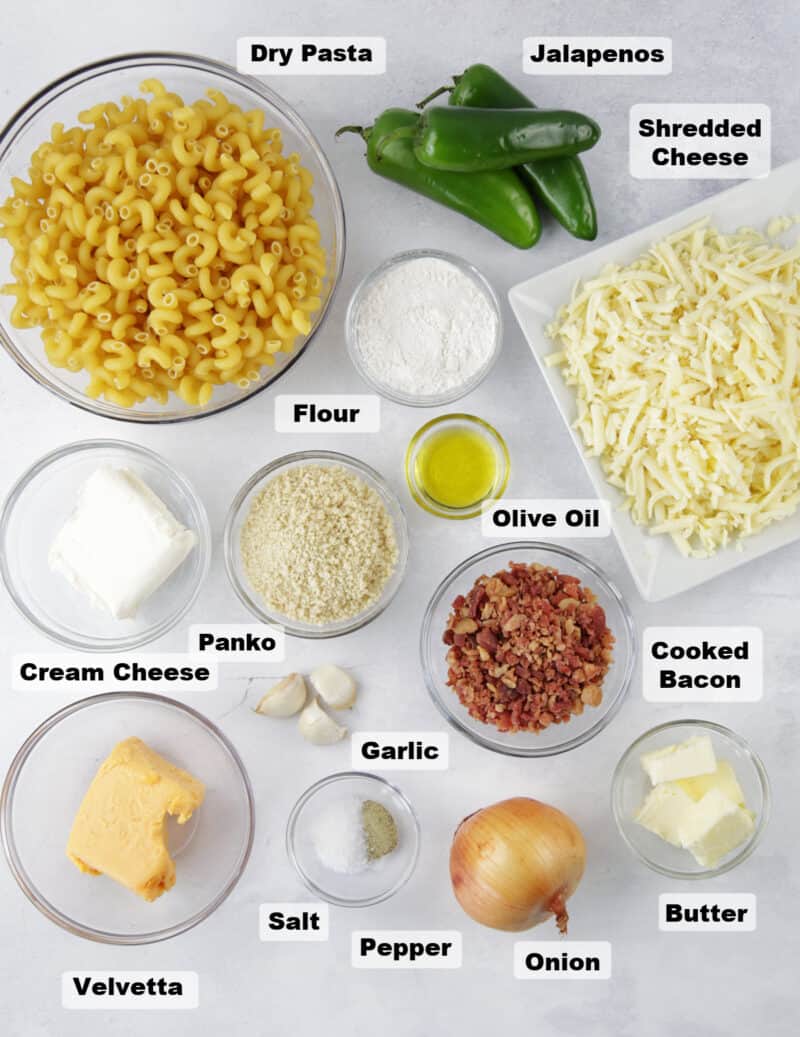 ingredients for Jalapeño Popper Mac and Cheese on a table including uncooked spiral noodles, shredded cheese, garlic cloves, a whole onion and fresh jalapeno poppers