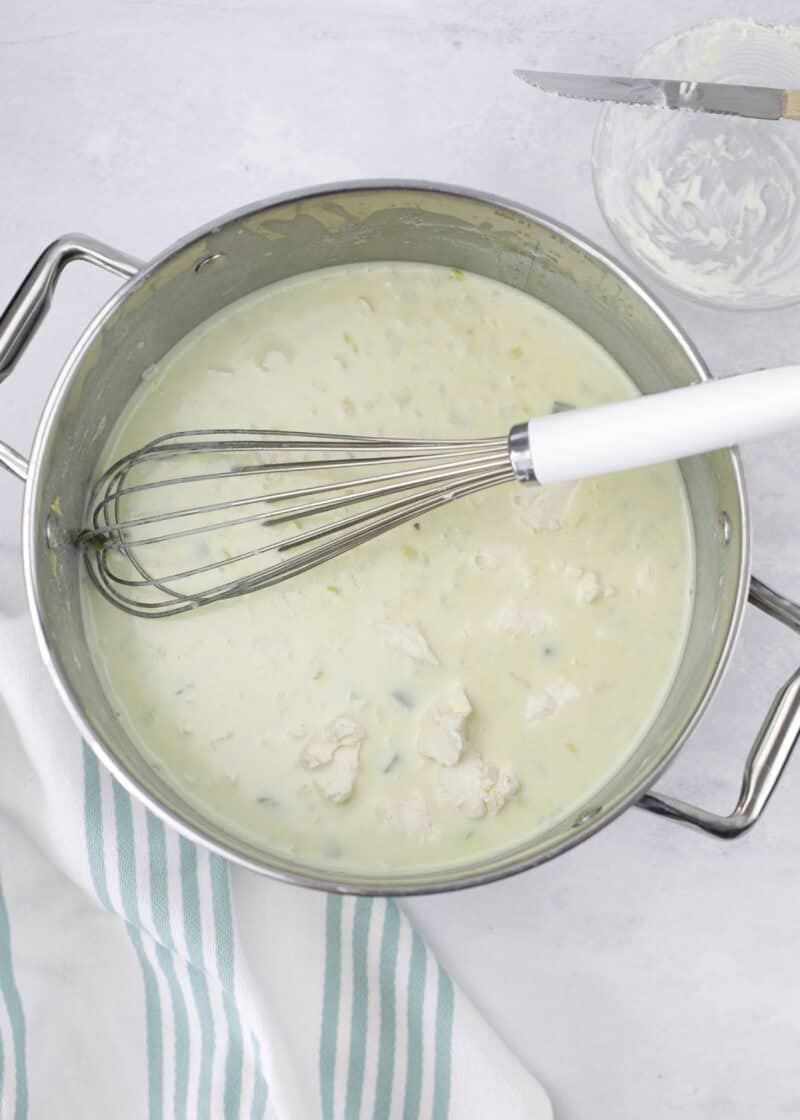 white sauce with chunks of cream cheese in a stainless steel pot on a table with a whisk