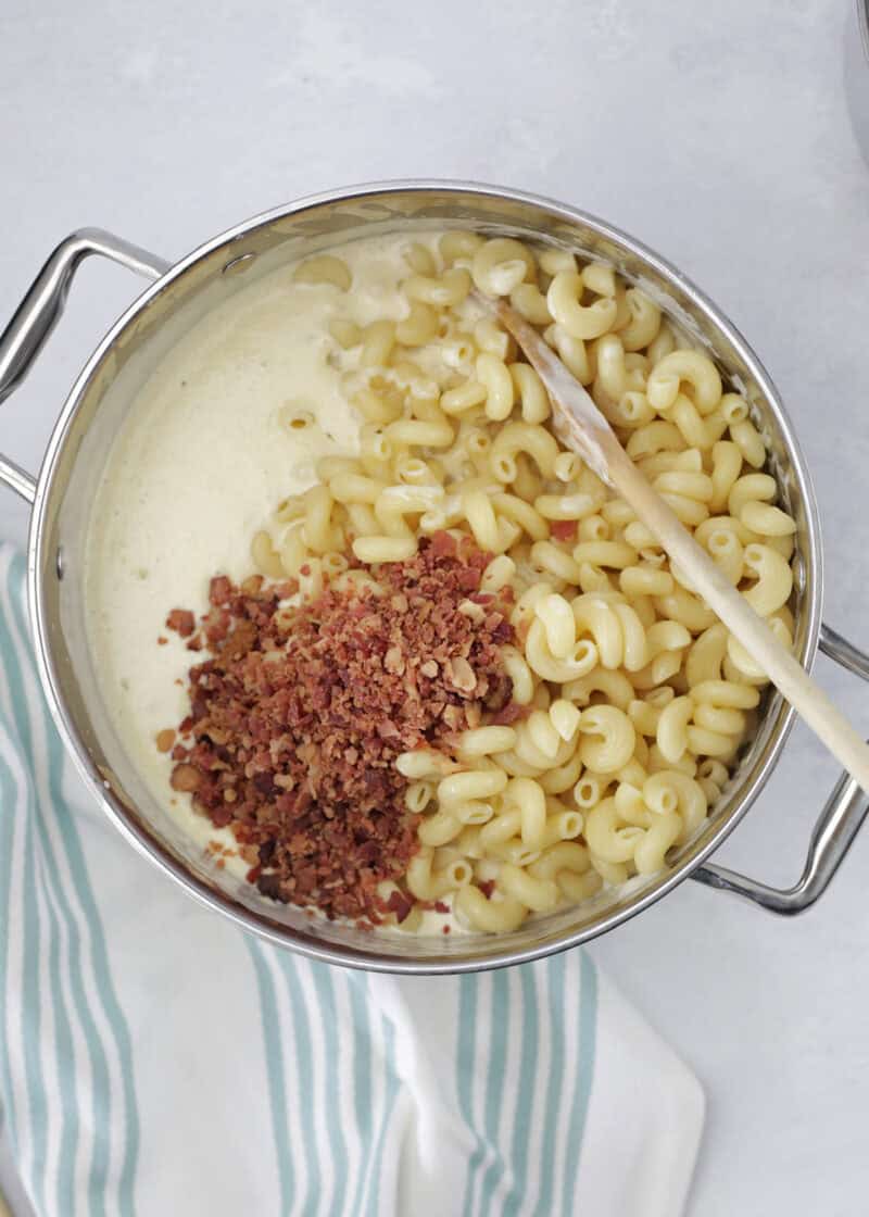cooked spiral noodles, cooked chopped bacon and a white sauce in a large stainless steel pot with a wooden spoon