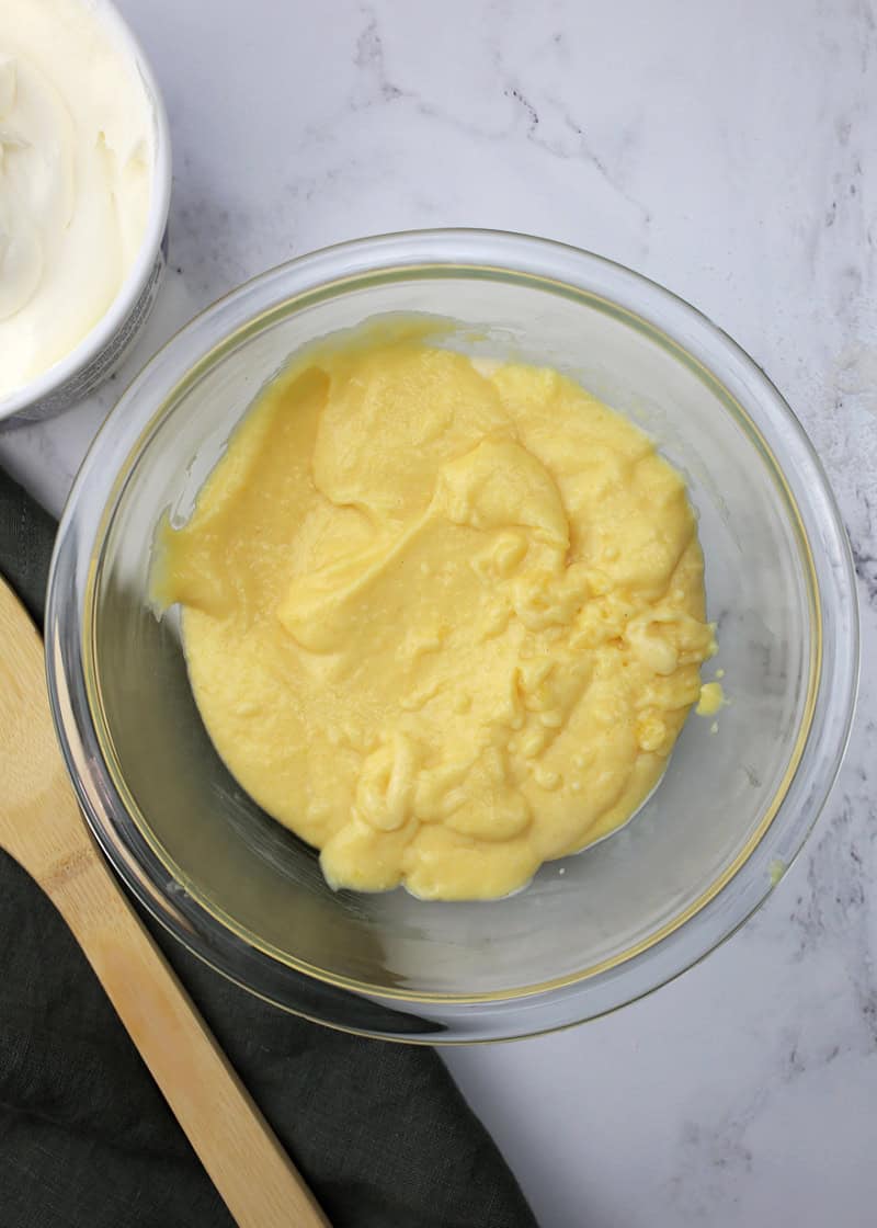 yellow pudding in a glass bowl with a wooden spoon beside it along with a tub of cool whip.
