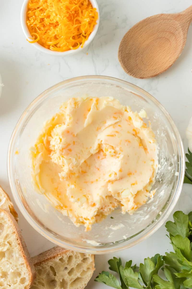 homemade garlic butter in a clear bowl on a table next to a bowl of shredded cheese and fresh sliced bread.