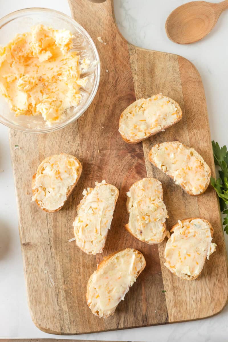 slices of bread with garlic butter and shredded cheese on it, on top of a wooden cutting board beside a bowl of cheesy garlic butter.