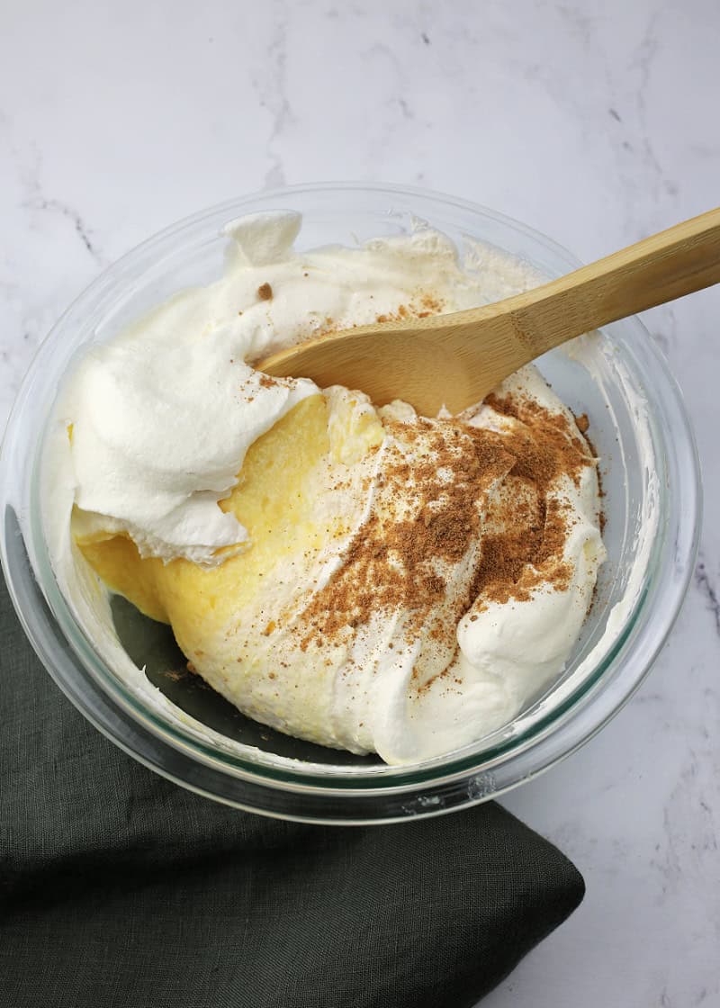 pudding, whipped cream, and nutmeg being mixed together in a glass bowl with a wooden spoon.