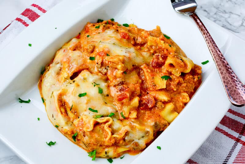 lasagna on a white plate with fresh parsley sprinkled on top, and a fork beside it.