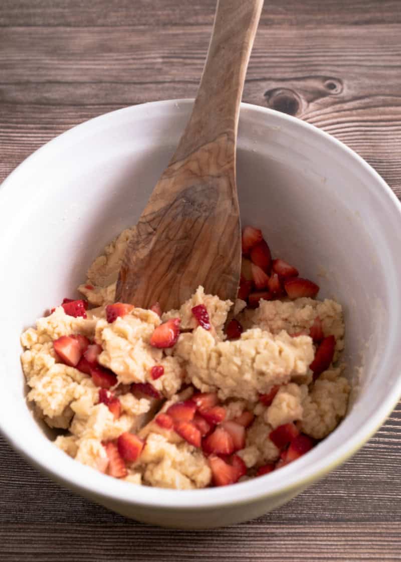 a white bowl with an uncooked cookie dough mixture with strawberry pieces in it with a wooden spoon.