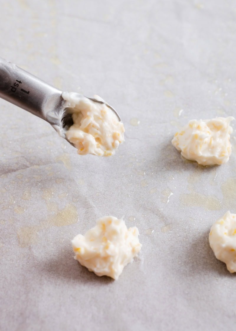 lemon cream cheese mixture being dropped onto parchment paper using a teaspoon measuring spoon.