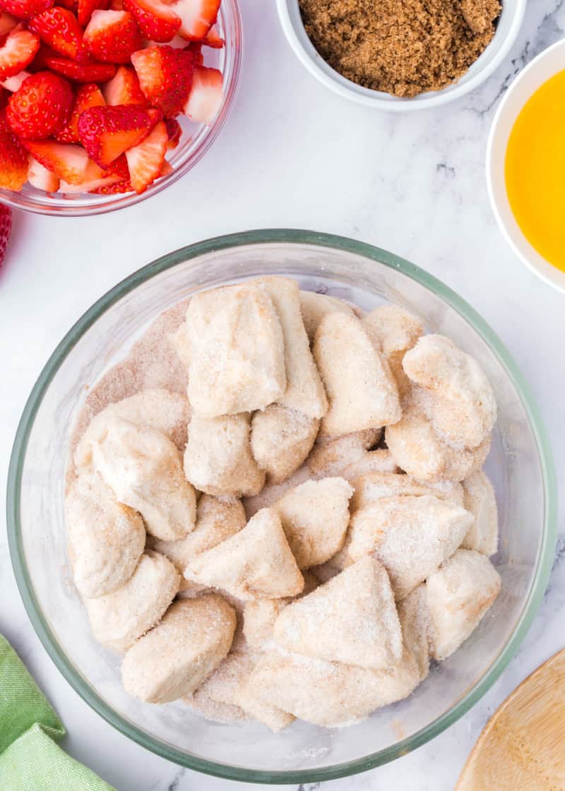 a clear bowl of uncooked cubes of dough tossed in a sugar mixture beside bowls of fresh cut strawberries and brown sugar.