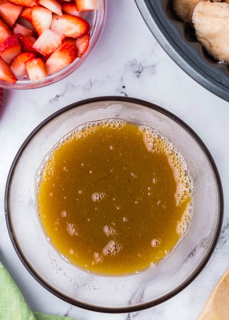 a clear bowl of melted butter and brown sugar beside a clear bowl of cut fresh strawberries and a bundt pan.