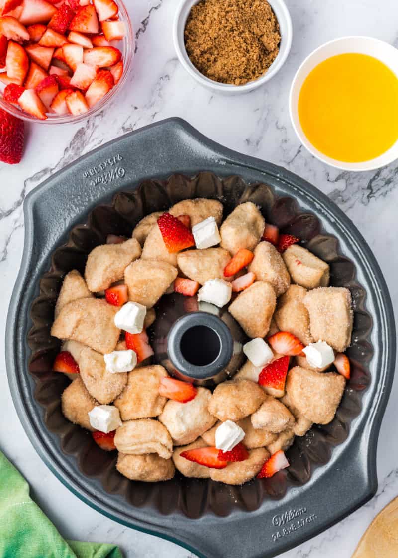 uncooked monkey bread with cubes of cream cheese and fresh cut strawberries in a bundt pan beside a bowl of brown sugar and cut fresh strawberries.