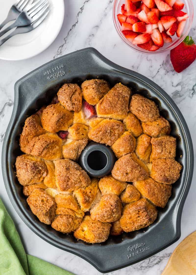 cooked monkey bread in a bundt pan on a table beside plates and a bowl of cut fresh strawberries.
