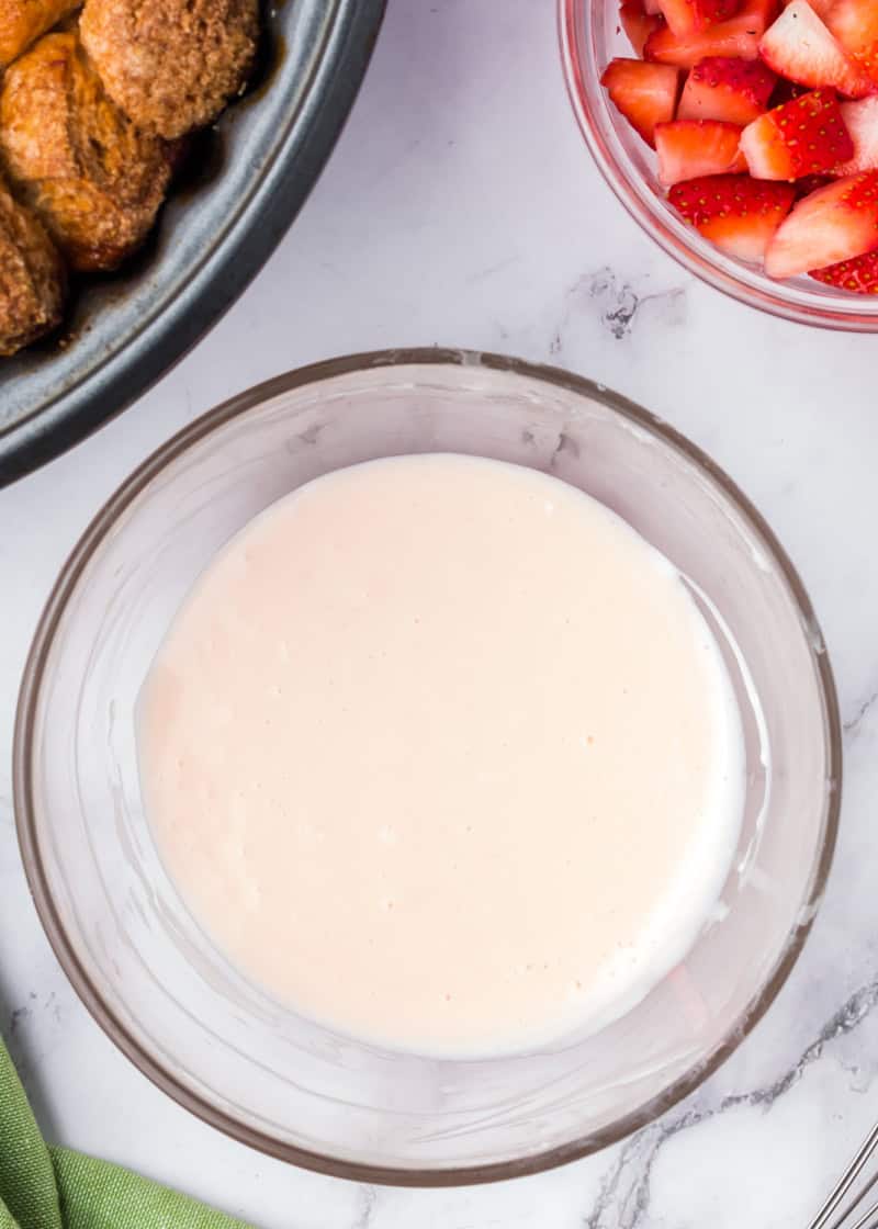 a clear bowl of white glaze on a table beside cut fresh strawberries and monkey bread.