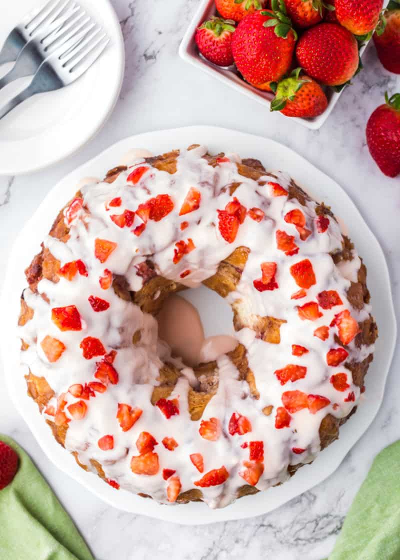 overhead view of a whole strawberry monkey bread in a bundt form with white glaze and strawberry pieces on it, surrounded by plates and a basket of strawberries.