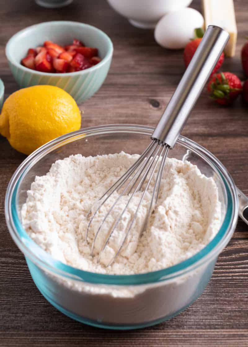 a glass bowl of a flour mixture with a metal whisk in it on a table with a whole lemon and a bowl of cut strawberries.