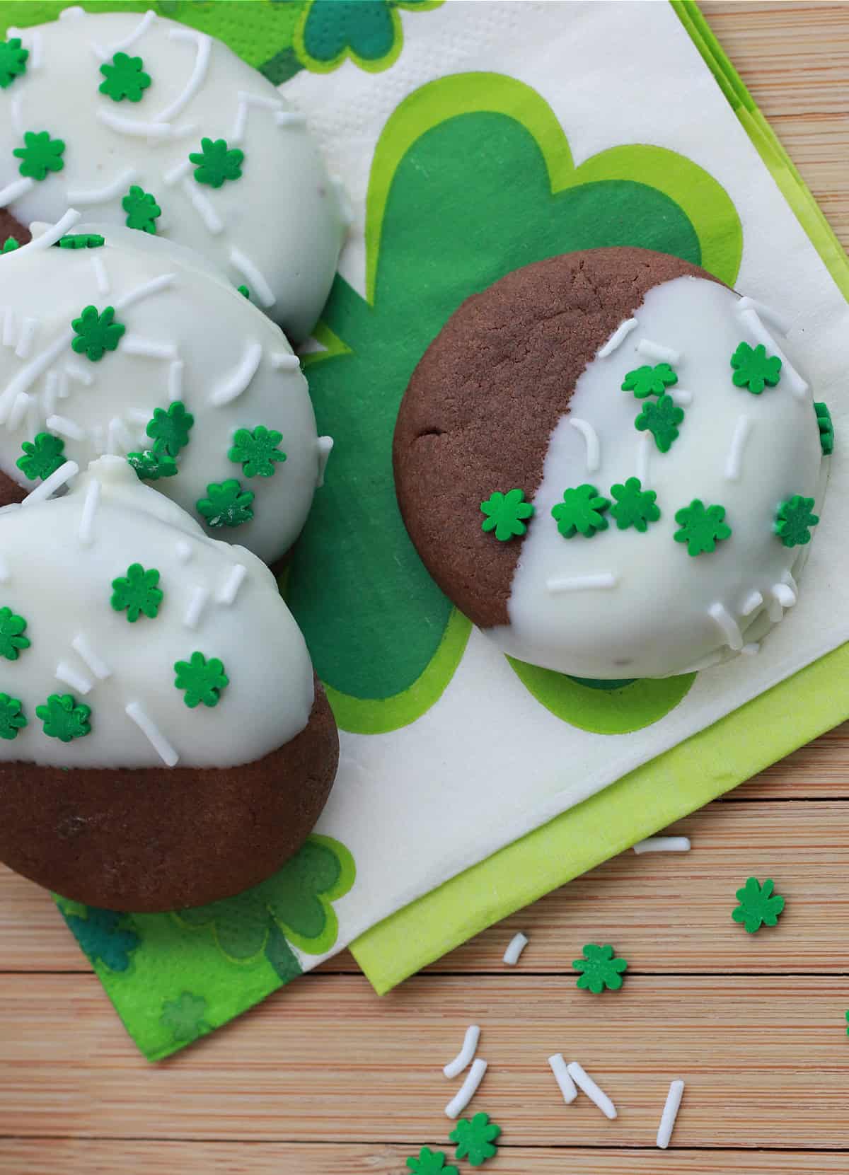 chocolate cookies half dipped in white chocolate with clover leaf sprinkles on a napkin.
