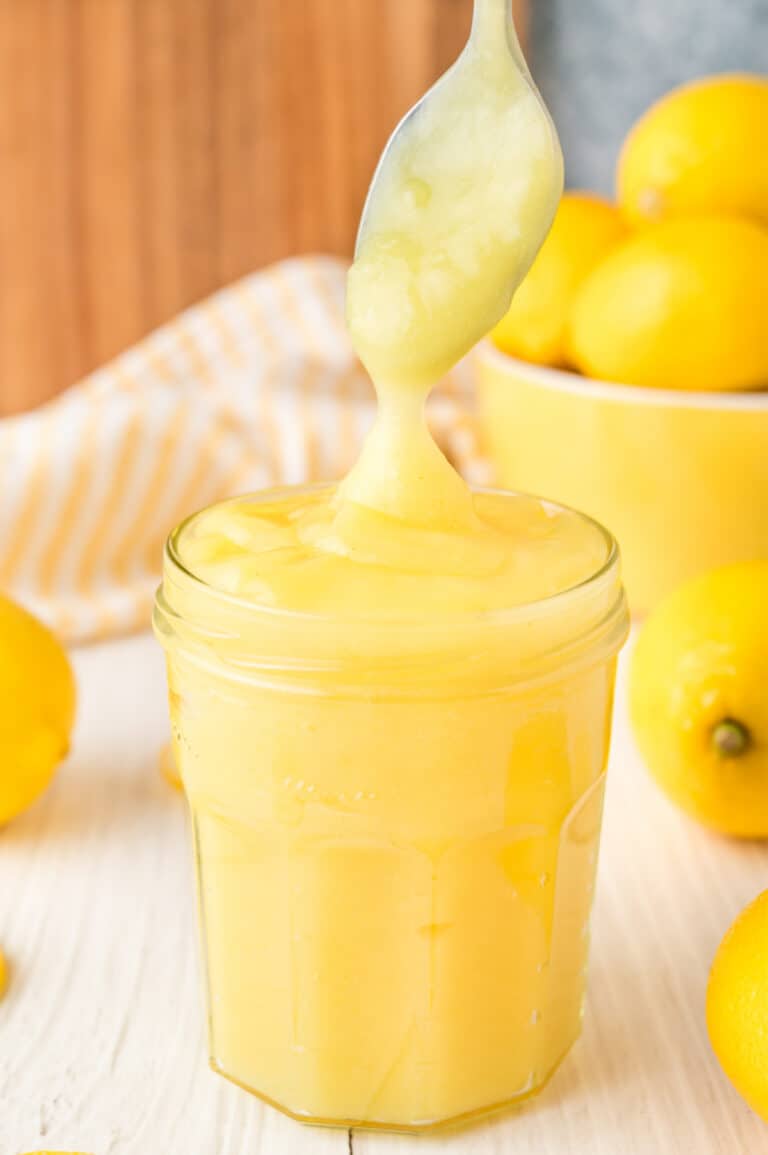 cooked bright yellow lemon curd in a jar with a spoon taking some out with fresh lemons behind the jar.