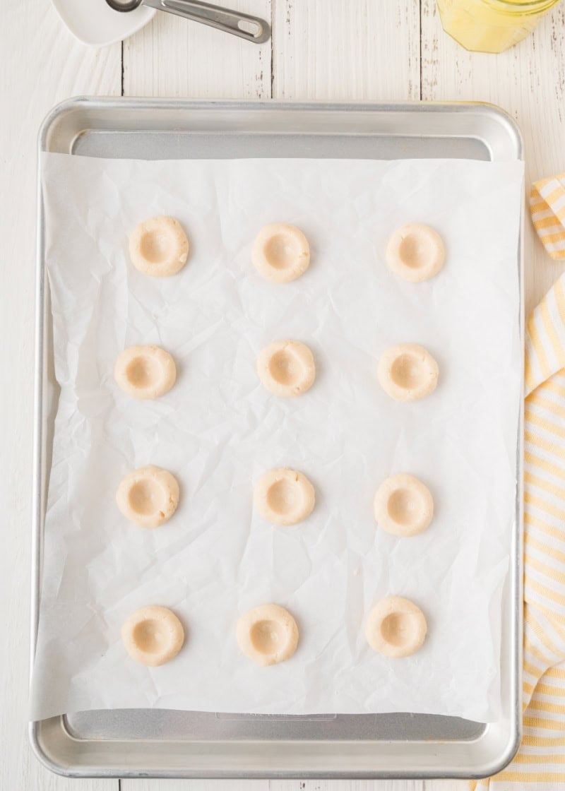 uncooked cookies on a parchment lined baking pan with a hole indented into each one for filling to go into.