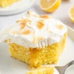 A white round plate with a slice of lemon poke cake with toasted meringue on top, with some cake on a fork beside it.