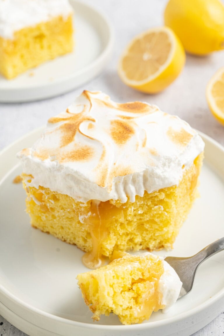 A white round plate with a slice of lemon poke cake with toasted meringue on top, with some cake on a fork beside it.