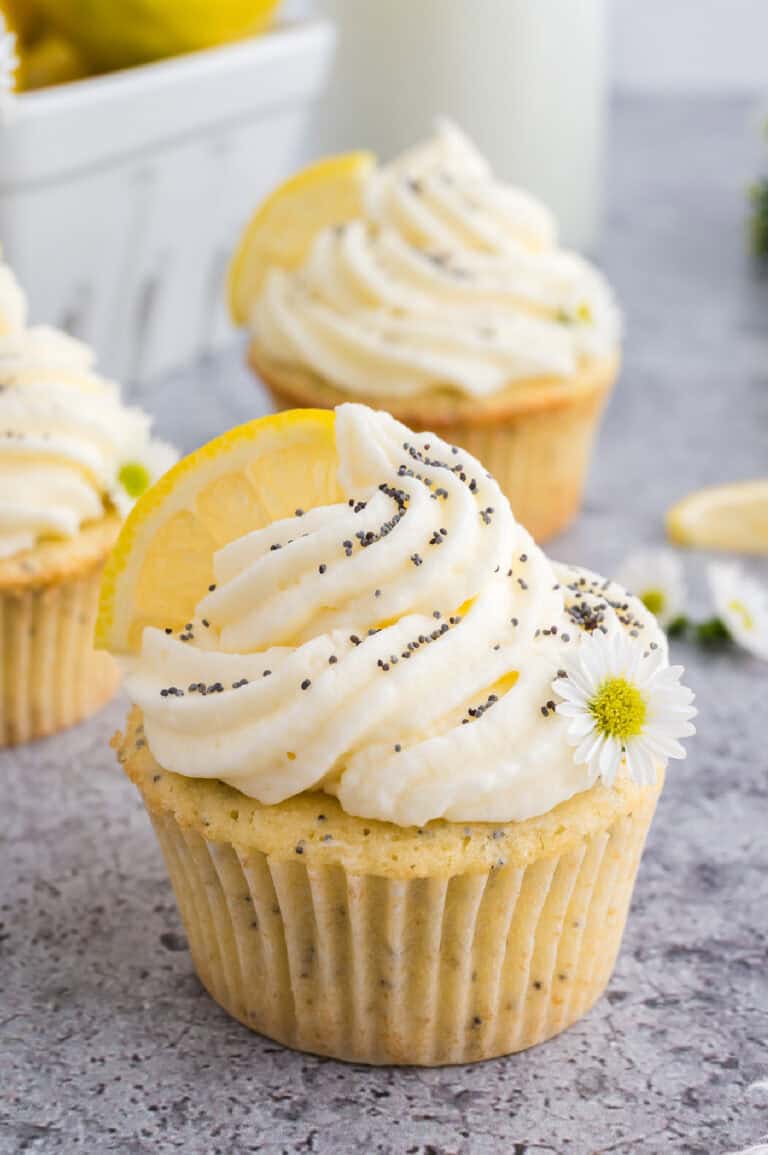 Lemon poppyseed cupcakes with a white buttercream icing on top, sprinkled with poppy seeds and a fresh lemon wedge on top.