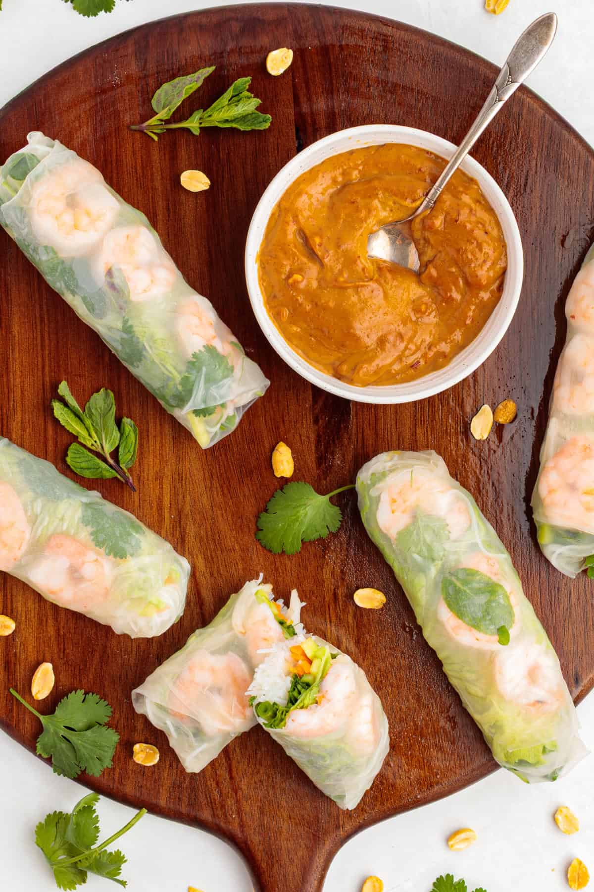 Vietnamese Shrimp Salad Rolls on a white plate with fresh herbs around it and a small bowl of peanut dipping sauce.