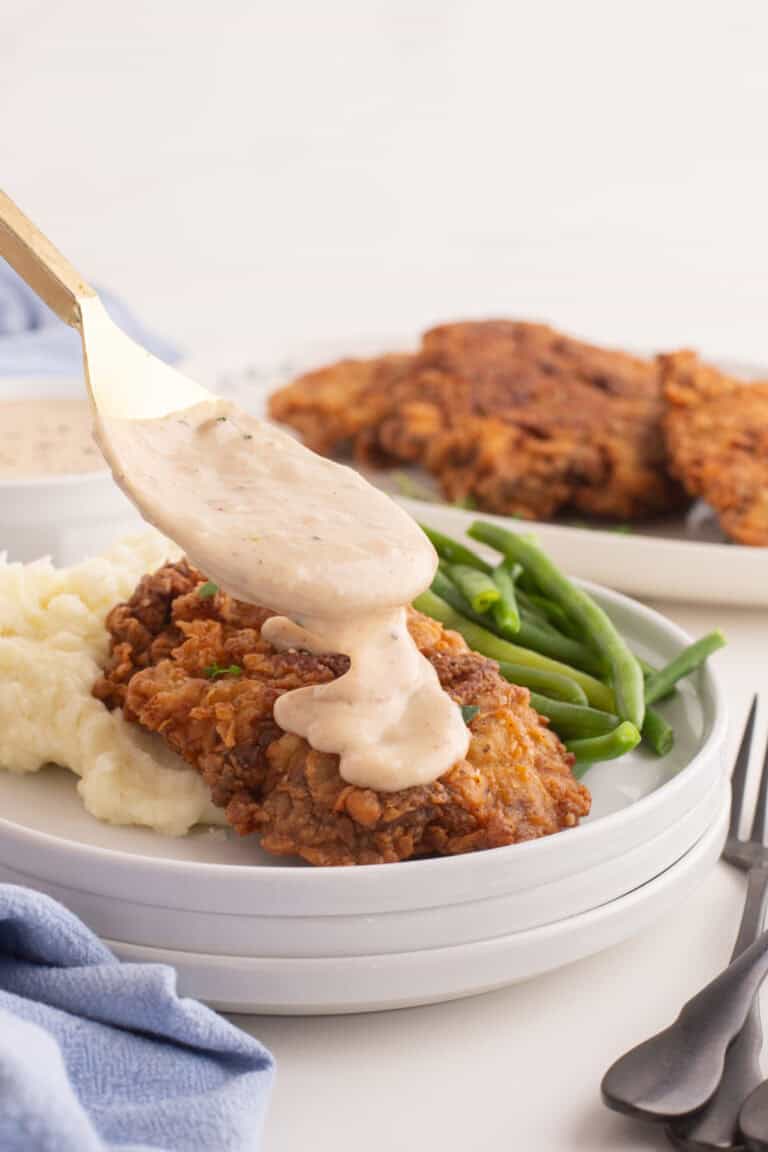 white gravy being pored onto chicken fried steak on a plate with mashed potatoes and green beans.