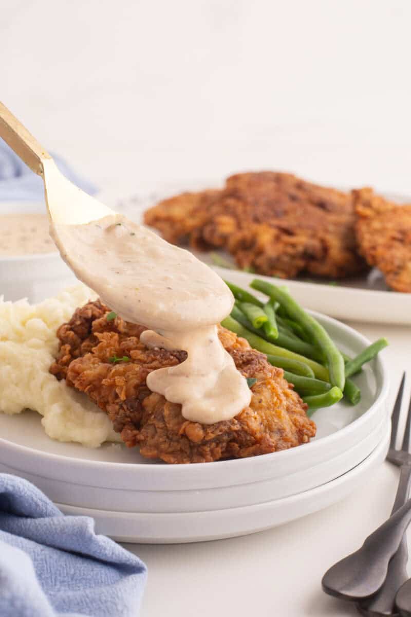 white gravy being pored onto chicken fried steak on a plate with mashed potatoes and green beans.