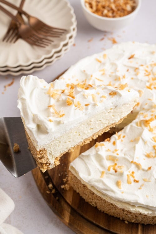 A slice of coconut cheesecake being taken out of a whole cheesecake sitting on a wooden plate.