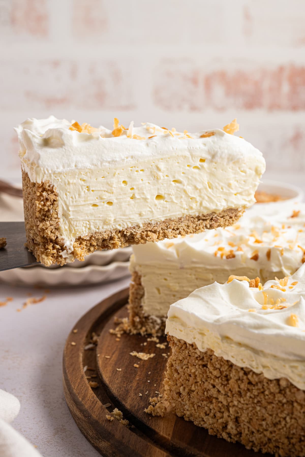 A slice of coconut cheesecake being taken out of a whole cheesecake sitting on a wooden plate.