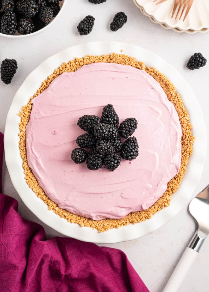 A whole Blackberry Cream Pie in a graham cracker crust with fresh blackberries on top, surrounded by fresh blackberries and plates.