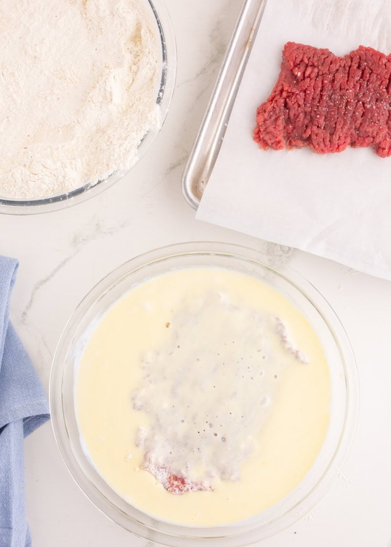 a piece of cube steak in a glass bowl with an egg mixture inside, and a bowl of flour next to it.