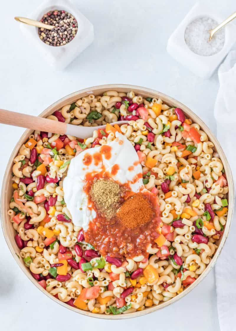 Mexican Macaroni Salad with sour cream and spices being mixed in, with a wooden spoon in it.